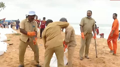 Sanitation workers on a beach filling sandbags, wearing uniforms, gloves, and masks, with others working near the shoreline.