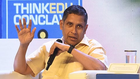 Image shows Praveen Chakravarty seated on a white chair, holding a microphone in one hand and gesturing with the other during a panel discussion. He is wearing a pale yellow shirt and a black wristwatch. Behind him is a blue sign that reads “THINKEDU CONCLAVE.” A placard on the table in front of him displays the name "SASTRA" — a reference to SASTRA Deemed University. The setting suggests a formal or academic event.
