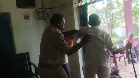 A police officer is seen restraining a man in a white shirt while another man stands nearby, gesturing. The scene appears tense and takes place indoors, possibly in an office or government facility, with a turquoise curtain and wall-mounted devices in the background.