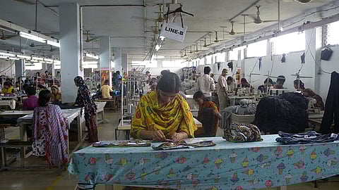 A large garment factory floor filled with rows of women workers, mostly dressed in colorful traditional attire, sitting at sewing machines. In the foreground, one woman in a yellow scarf focuses on trimming fabric pieces on a table covered with a bright, cartoon-patterned cloth. The room is lit with fluorescent lights and ceiling fans, with a sign reading "LINE-B" hanging above the workstations.