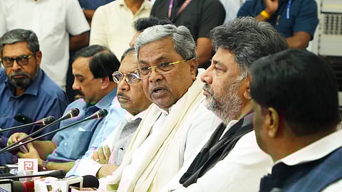 A group of Indian politicians is seen seated at a press conference. Karnataka Chief Minister Siddaramaiah is at the center, speaking into multiple microphones from various media outlets. Deputy Chief Minister D.K. Shivakumar sits beside him, along with other officials. The background shows more attendees and journalists, suggesting an important announcement or briefing.