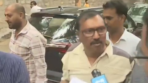 A group of men are gathered near a parked black vehicle on a street. The man in the center, wearing glasses and a beige shirt, is holding papers and speaking to a journalist who is holding a microphone labeled "NTV". Other men are seen standing around, and there are more vehicles and a stone wall in the background.








