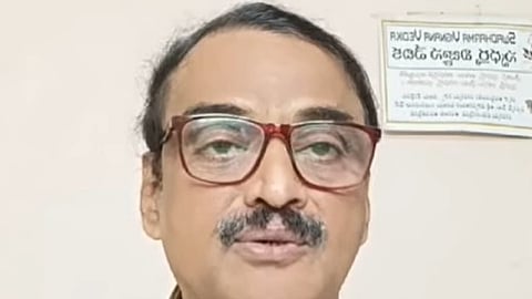 A middle-aged man with short black hair, a trimmed moustache, and large maroon-rimmed glasses is seen speaking. He is wearing a multicolored checkered shirt and is positioned against a plain indoor background with a Telugu signboard visible on the wall behind him.