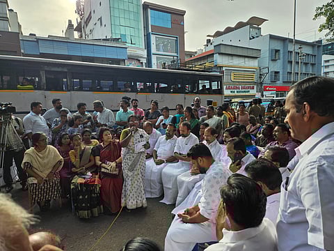 A large group of ASHA workers and their supporters sit and stand in protest on a busy street in front of the Kerala Secretariat. One woman speaks into a microphone, while others listen. Buses and buildings are visible in the background, with a crowd of people surrounding the seated protesters.
