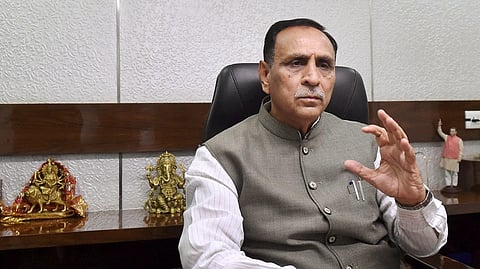 A man is seated at a desk in an office, gesturing with his right hand while speaking. He is wearing a grey Nehru jacket over a white shirt and has pens in his chest pocket. Behind him, there are small statues of Hindu deities including Durga and Ganesha, as well as a figurine of another man with one arm raised. The setting appears formal, suggesting an official or political environment.