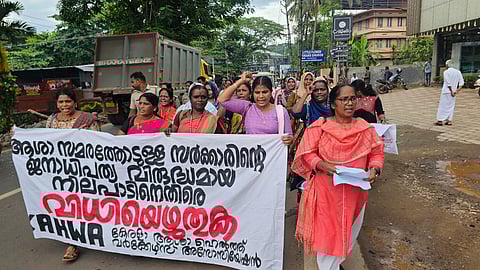 A group of ASHA workers march on a road holding a banner written in Malayalam, protesting government actions, with one woman leading the chant.
