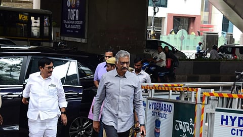 Two men, including a senior official in a grey shirt and dark trousers, are walking past police barricades marked "Hyderabad City Police" near the Jubilee Hills Law & Order Police Station. They appear to be arriving or departing from a location under a flyover, with security and media presence visible. A black vehicle is parked behind them, and a Telangana Gaddar Film Awards poster is displayed on a pillar in the background.