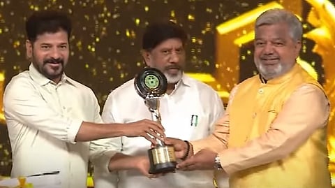 Three men are seen on stage during an award ceremony. The man on the left, wearing a white shirt, and the man on the right, dressed in a yellow kurta, are smiling as they hold a trophy together. A third man, also in a white shirt, stands in the middle behind them, observing the moment. The background is decorated with golden lights and patterns, giving a celebratory feel.