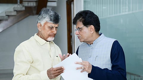 Two men are seen standing indoors, engaged in a serious conversation while looking at a document. The man on the left is wearing a light yellow shirt, and the man on the right is dressed in a blue kurta with a light grey checkered vest. A staircase and glass panel are visible in the background, suggesting a formal setting.