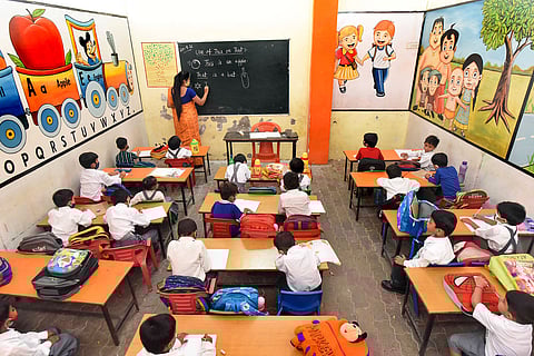 A brightly colored classroom filled with young children seated at desks, attentively watching a female teacher writing on a blackboard. The blackboard displays a lesson on "Use of This or That" with simple sentences like "This is an apple" and "That is a bat." The classroom walls are decorated with educational and cartoon-themed murals, including an alphabet train and cheerful children holding hands. The students, wearing school uniforms, are engaged with notebooks and school bags on their desks, creating a lively and vibrant learning environment. 