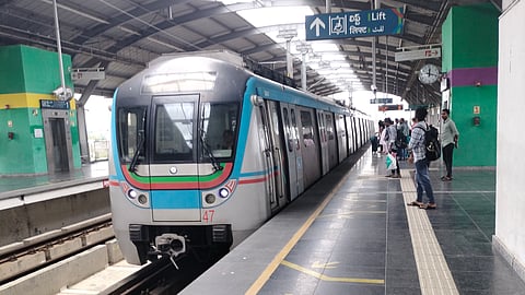 A Hyderabad Metro train arrives at an elevated station platform. Passengers wait near the edge, standing behind safety lines marked with directional arrows. The station has a covered roof, green and purple painted pillars, signage in multiple languages, and a visible clock and lift directions above.