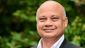 A bald man with light brown skin wearing a black suit jacket over a white shirt, smiling softly while standing outdoors against a background of green foliage. A small cross pin is visible on his lapel.
