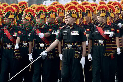 A group of Indian Army soldiers in ceremonial uniform march in formation, wearing red and yellow fan-shaped headgear and black outfits with striped sashes and white gloves. The officer at the front leads with a drawn sword.