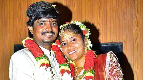 A newlywed couple dressed in traditional South Indian attire poses closely together, smiling. The groom wears a white sherwani and the bride is adorned in a red and cream saree with gold embroidery. Both wear garlands made of red, white, and yellow flowers and have ceremonial decorations like bangles, gold jewelry, and floral headpieces. They are holding hands affectionately against a wooden-panel background.