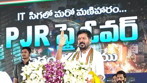 Telangana CM Revanth Reddy stands at a podium adorned with vibrant floral arrangements as he addresses a public gathering. Behind him is a large digital display with bold Telugu and English text reading "PJR Flyover" and "IT సిటీలో మరో మణిహారం…" ("Another jewel in the IT city..."). The background features an illuminated urban cityscape, suggesting a development project announcement or inauguration. The man raises one hand in a gesture while wearing a white shirt and a tricolor scarf, indicative of Indian political symbolism. 
