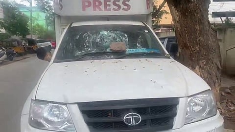 A white Tata vehicle with a “PRESS” board mounted on top is parked beside a tree. The windshield is severely damaged, with a large brick resting on the hood. Debris and leaves are scattered across the vehicle’s front, and a blue Mahaa News banner is visible inside the windshield. The vehicle appears to have been targeted in an act of vandalism.