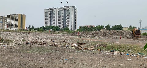 A large open area filled with legacy waste and debris from illegal dumping at Pallavaram Periya Eri, with high-rise apartment buildings in the background. Piles of garbage, a cement mixer, and makeshift barriers are visible, highlighting environmental degradation amid urban development.