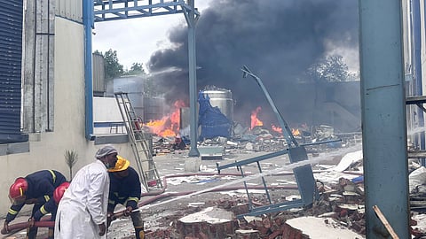 A large industrial fire is seen raging at a factory site, with thick black smoke and flames rising from the debris. Several firefighters in protective gear are on the scene, unrolling hoses to control the blaze. A man in a white coat stands nearby, possibly a worker or supervisor. The ground is covered in rubble and twisted metal, indicating a possible explosion or structural collapse. A large silver tank and partially destroyed equipment are visible amid the destruction.