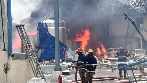 Firefighters battle a massive industrial blaze amid thick black smoke and flames in Sangareddy. The scene is chaotic, with debris scattered across the ground, a metal tank partially covered in blue plastic, and a burnt-out car engulfed in fire. Three firefighters in protective gear and helmets are seen handling a fire hose in the foreground while another walks nearby. The aftermath of the explosion has left the area in ruins, with collapsed structures and broken materials visible throughout.