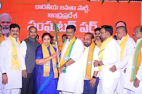 A group of Bharatiya Janata Party (BJP) leaders and members are seen on stage at a political event in Andhra Pradesh. A woman in a blue saree, wearing a BJP scarf, hands over a party flag to a man in white traditional attire, also wearing a BJP scarf. Other leaders stand alongside them, all dressed in white with orange BJP scarves, against a bright orange backdrop displaying the party's name in Telugu. Portraits of national BJP leaders, including Prime Minister Narendra Modi, are partially visible in the background. The atmosphere appears celebratory and symbolic of new membership or alliance.