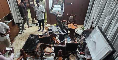 A room in severe disarray, with broken furniture, overturned chairs, shattered glass, and a refrigerator lying open and toppled on its side. Several men stand among the debris, observing the damage. The floor is littered with glass shards, food, and scattered household items. 