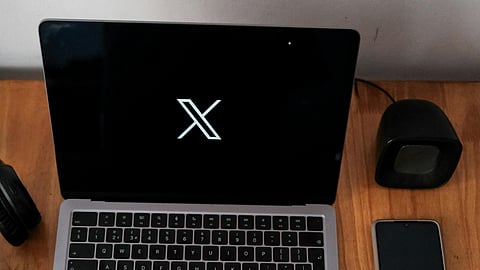 A laptop and smartphone placed on a wooden table, both displaying the white "X" logo on a black screen, representing the rebranded Twitter (now X). A small black speaker is positioned next to the phone.