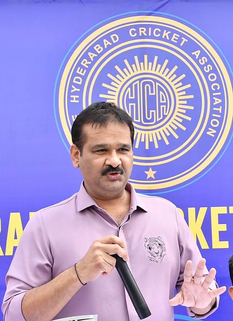 A man in a light purple shirt with a tiger emblem is speaking into a microphone at a public event. He stands in front of a blue and yellow backdrop featuring the logo of the Hyderabad Cricket Association (HCA), with "HCA" in the center and the full name encircling it.
