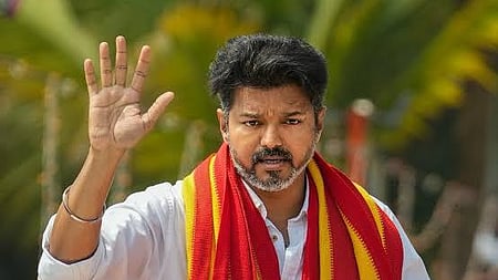 A medium-close-up photo of Indian actor and politician Vijay. He is looking slightly to his left and has a serious expression. He is wearing a white shirt and is draped in a thick red and yellow striped shawl (or anga vastram), which is likely associated with his political party, Tamilaga Vettri Kazhagam. His right hand is raised in a gesture that resembles a salute or a wave. He has a neatly trimmed beard and mustache. The background is slightly blurred and features green foliage and what appears to be a light-colored structure.