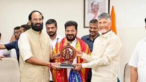 Telangana Chief Minister Revanth Reddy, flanked by Andhra Pradesh Chief Minister N. Chandrababu Naidu and Telangana minister Uttam Kumar Reddy, smiles while receiving a decorative idol as a gift during a meeting. All three leaders are holding the idol together. Revanth is draped in a ceremonial shawl. Several people and an Indian flag are visible in the background, along with a framed portrait of President Droupadi Murmu on the wall.