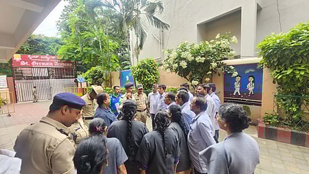 A group of police officers and civilians gather inside the premises of St. Germain Academy in Bengaluru. The scene appears tense, with officers speaking to a group of concerned individuals, possibly school staff or parents. The school entrance is visible in the background, with murals of children on the walls and greenery surrounding the courtyard.
