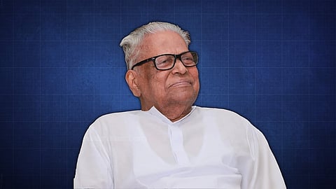 Portrait of veteran Communist leader VS Achuthanandan wearing a white shirt and black-rimmed glasses, smiling gently, set against a blue background with a subtle grid pattern.