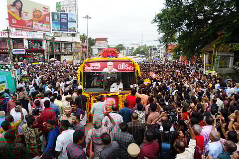 A large crowd gathers on both sides of a street in Kerala as a flower-decorated bus carrying the body of V S Achuthanandan moves slowly during his funeral procession, with people taking photos, holding banners, and police managing the crowd.