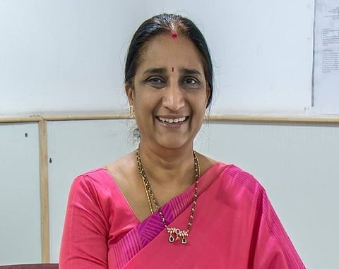 A middle‑aged woman wearing a pink saree and gold jewellery, smiling at the camera in an indoor setting.