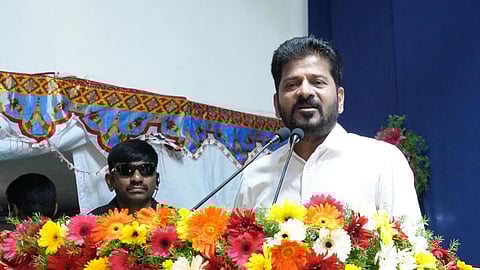 Revanth Reddy in a white shirt is speaking at a podium decorated with a vibrant arrangement of red, yellow, orange, and white flowers. He is flanked by two microphones. Behind him, a man in black sunglasses and a headset appears to be providing security. The background includes a blue curtain and a tent-like decorative canopy with colorful patterns.