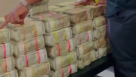 A man in a black shirt is standing beside a table stacked high with bundles of Indian currency notes, held together with colored bands. He appears to be pointing at one of the stacks, while resting his other hand on the money. The scene is indoors, with green and patterned curtains visible in the background.
