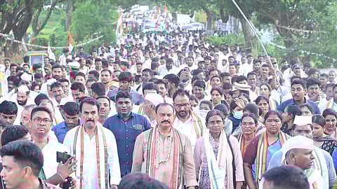A large crowd of people participates in a political padayatra (foot march) on a road lined with trees and Indian national flags. Many individuals in the front wear white clothes and tricolor scarves, indicating affiliation with the Indian National Congress party. Some leaders are centrally positioned, walking at the head of the group. The atmosphere is energetic and peaceful, with people walking in close formation.