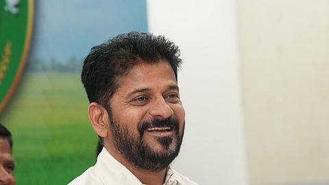 Revanth Reddy with a trimmed beard and short black hair is smiling while seated at a desk with papers and pens in front of him. He is wearing a cream-colored shirt and holding a paper and pen in his hands. A microphone is positioned near him, and there is a visible sign reading "I & PR" indicating an official press or public relations setting. The background shows a green field image and a blurred person sitting nearby.