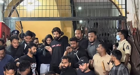 Actor Allu Arjun is surrounded by a large crowd of fans, security personnel, and police officers at the entrance of a theater. He is wearing a black t-shirt with red text and being escorted through the crowd. The setting appears to be a public event with tight security and significant attention.