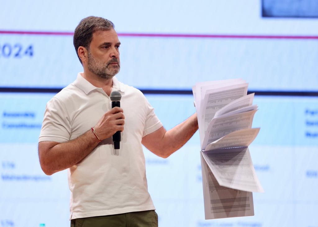 Rahulk Gandhi in a white tshort. Holding a think bunch of papers in one hand and a mic in the other