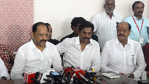 A group of men seated at a press conference table, with multiple microphones from various media outlets in front of them. Three men in white shirts are seated at the center, with others standing in the background, and a patterned wall partially visible on the left side.