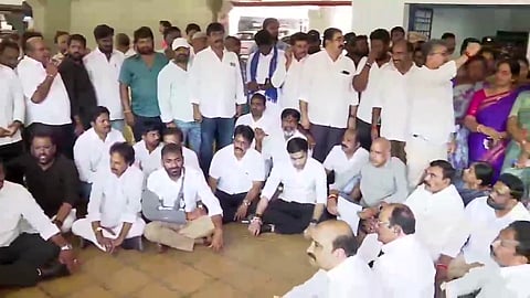 A large group of people, mostly men dressed in white shirts, are gathered indoors, with some sitting cross-legged on the floor and others standing behind them. A few individuals wear colored clothing, and one woman in a purple saree stands to the right. The group appears to be engaged in a meeting or protest, with attentive and serious expressions.