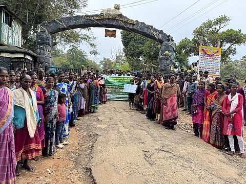 Men and women stand gathered on either side of a protest banner, in front of an arch that says, 'Nagarahole Tiger Reserve'. 
