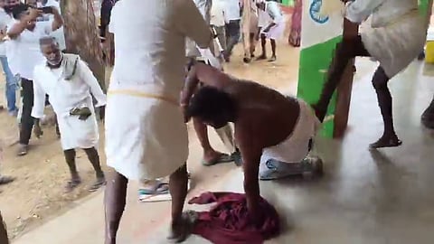 A man wearing a white dhoti sits on the ground near a pillar, with a maroon cloth in front of him, as another man in a white shirt and dhoti extends his arms nearby; several other people are seen standing in the background in an outdoor setting. 