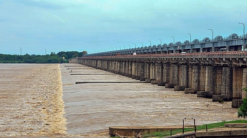 A wide view of Dowleswaram Barrage on the Godavari River, showing strong water flow beneath the long bridge-like structure with multiple pillars and gates. The cloudy sky and greenery in the background add to the scene, while vehicles can be seen moving on the road atop the barrage.