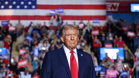 Donald Trump in a dark blue suit with a red tie stands in front of a large American flag. He is facing forward with a serious expression. Behind him, a large crowd is holding up red, white, and blue signs at what appears to be a political rally.