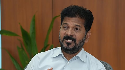 A man with a beard and short black hair, dressed in a light blue shirt, is seated at a desk with papers, files, and water bottles. He gestures with his hand while speaking, with a microphone placed in front of him. A green plant and wooden wall panels are visible in the background.