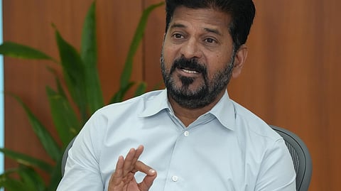 A man with a beard and short black hair, dressed in a light blue shirt, is seated at a desk with papers, files, and water bottles. He gestures with his hand while speaking, with a microphone placed in front of him. A green plant and wooden wall panels are visible in the background.