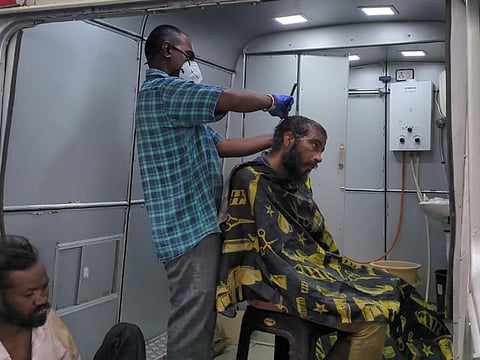 A man is receiving a haircut inside a mobile unit. The barber, wearing a face mask, gloves, and a blue checkered shirt, trims the hair of a seated man draped in a black and yellow cape. Another man, with unkempt hair and wearing a beige shirt, sits nearby, waiting. The setting is a service van with a sink and utility equipment visible in the background.
