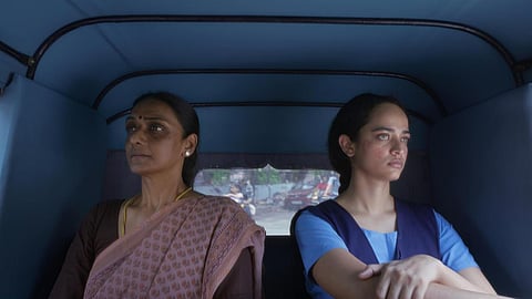 A older woman in a brown saree and blouse, and a young woman in a blue school uniform sit in an auto facing away from each other. 