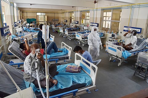 A crowded hospital ward is shown with many patients on beds. Most are elderly men, and some are connected to ventilators. A few healthcare workers in personal protective equipment (PPE) are attending to the patients. The room has yellow and white walls, and a large window is visible in the background.